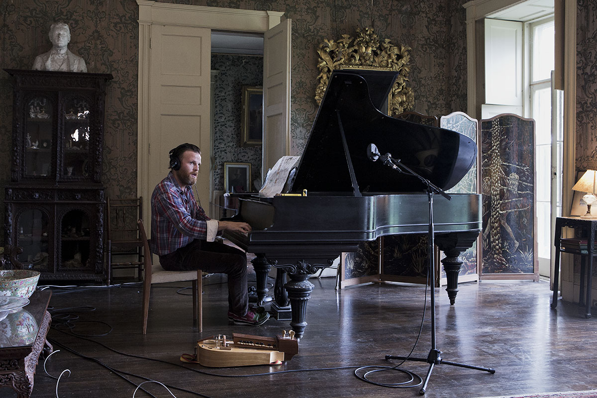 Photograph of a man playing a grand piano in a large room, a hurdy-gurdy sits at his feet 