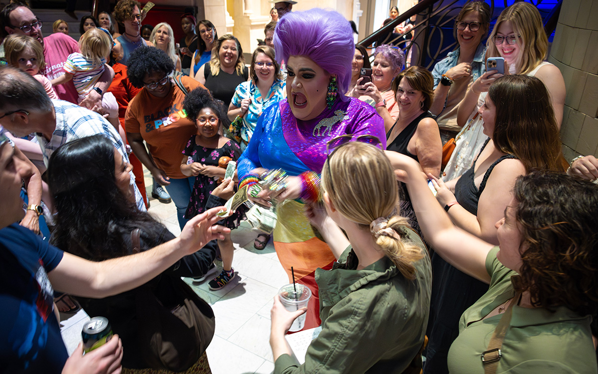 A white drag queen with big purple hair exclaims as visitors tip her cash