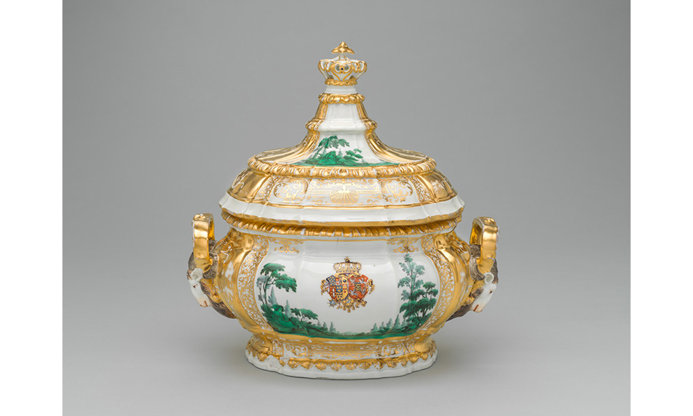 Tureen with Lid, a porcelain pot with gold trim, rams heads for handles, and a green forest scene with a royal coat of arms painted on the side. The lid has a tall crown handle with matching gold trim and green trees