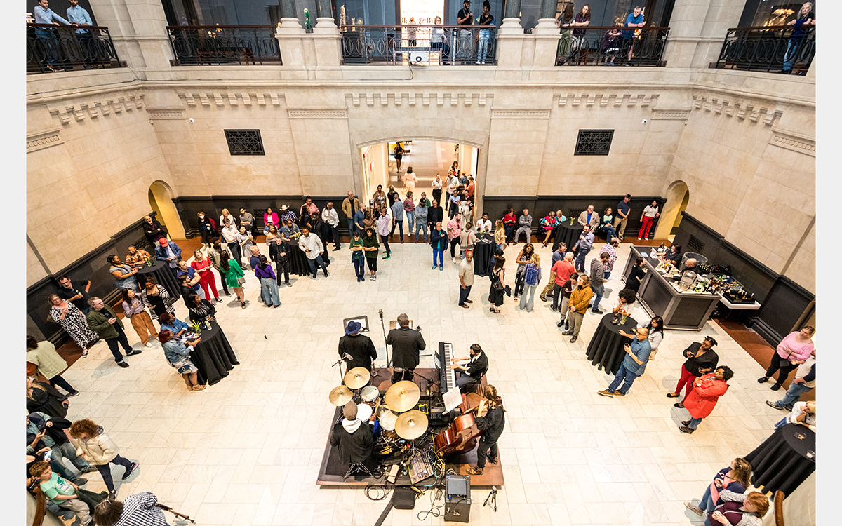 Above view of a two-story atrium with people standing at high-top tables and a band on a square stage in the center.