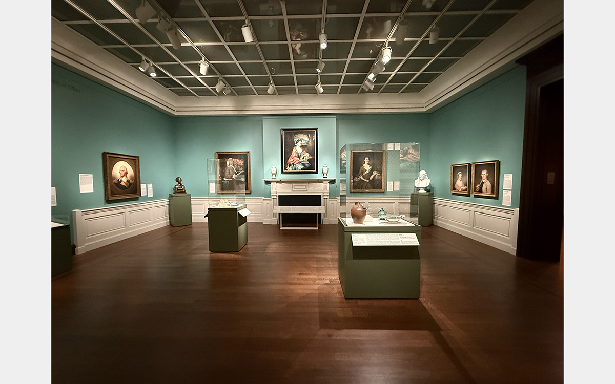 Museum exhibition gallery featuring a fireplace mantle, paintings, sculpture busts, and display cases.