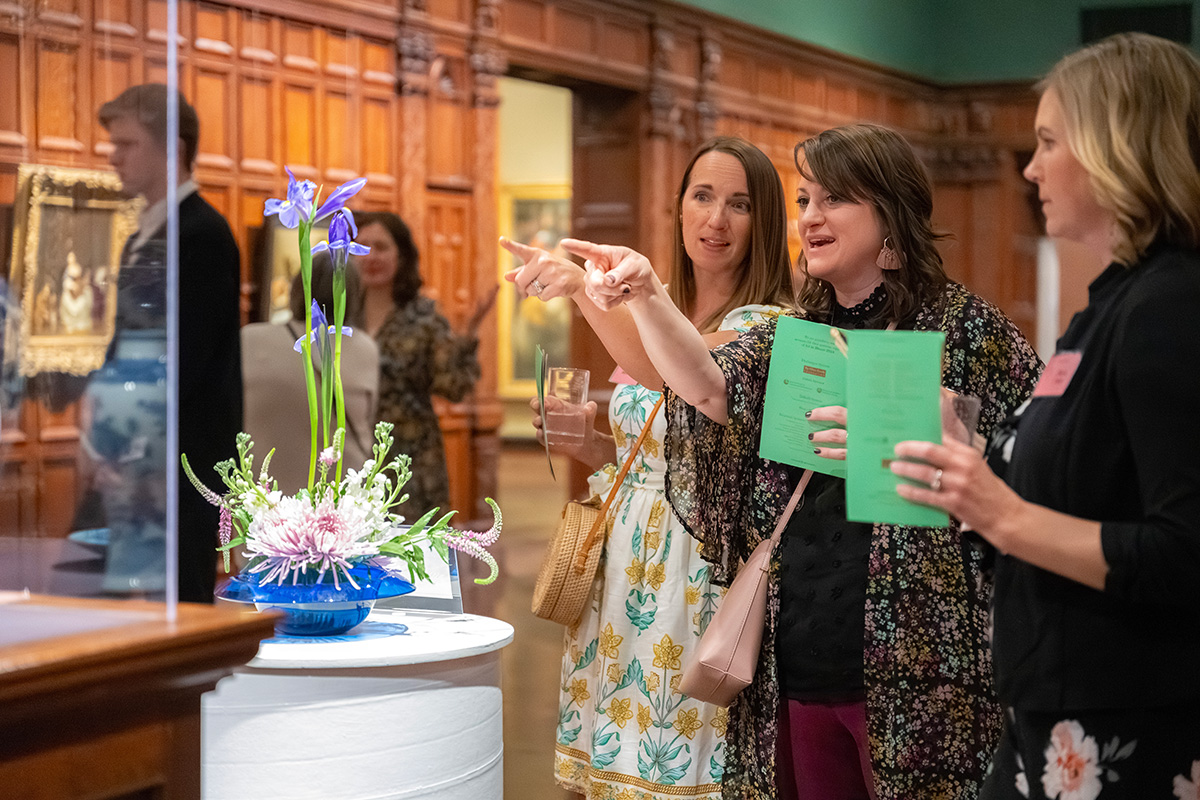 Three white women point at a floral arrangement in a gallery