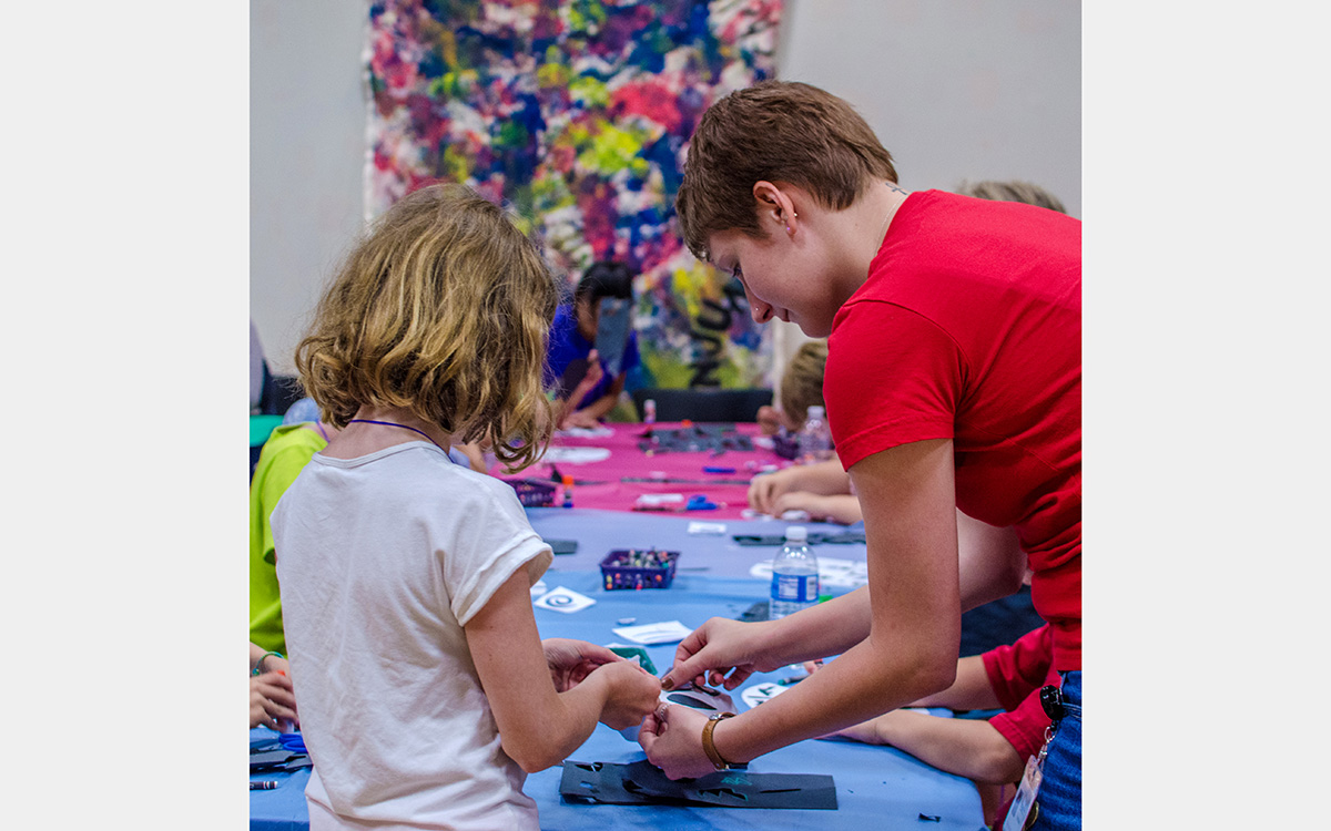 Girl and woman make crafts on a long table with a colorful painting in the background
