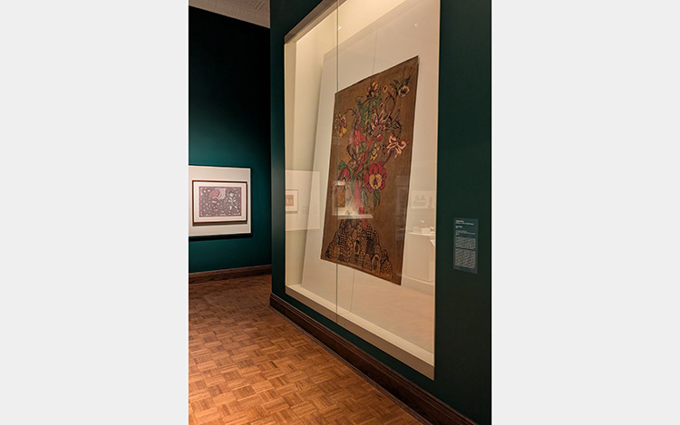Green-walled gallery space with wood parquet floor. Large, floor to ceiling display case with flat artwork featuring a colorful floral design on a brown background.