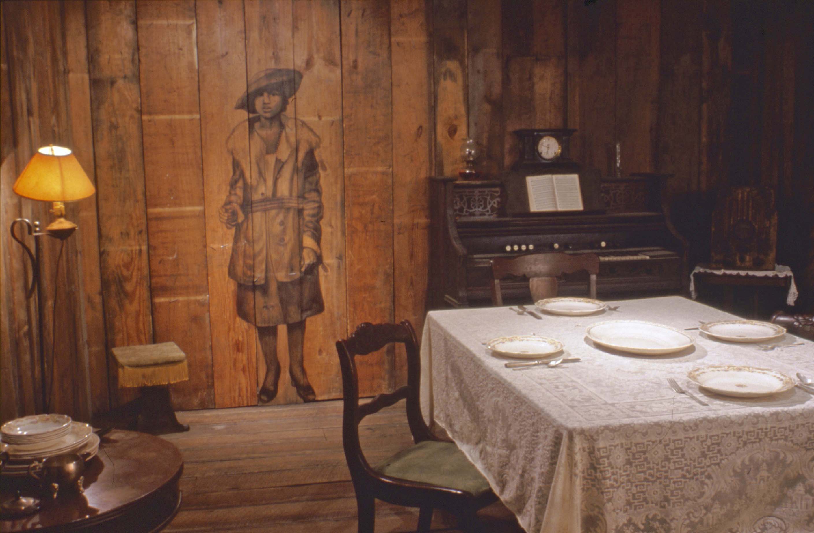 Installation picture of the work Visitation that includes a dining table, organ, and wooden walls.