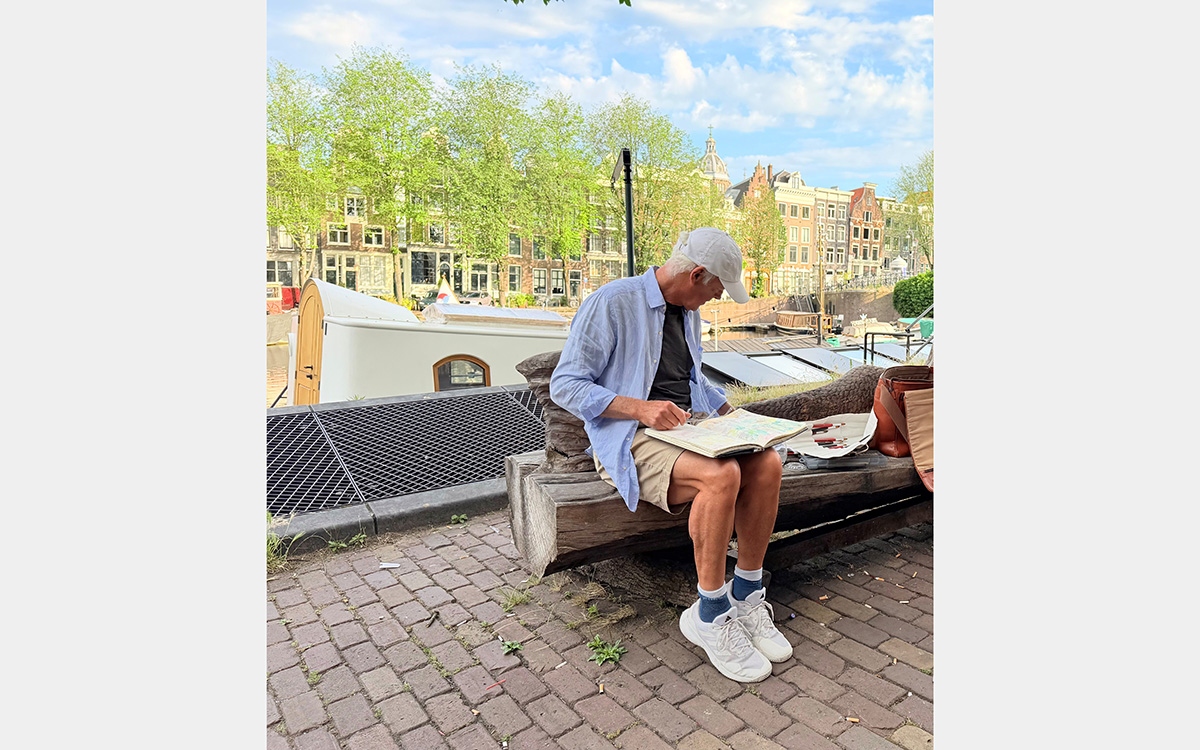 Man in shorts and ball cap sits on bench with sketchbook on lap against the backdrop of townhouses, tees, and boats.