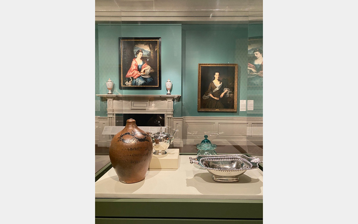 An image of a gallery in the museum, with walls painted in a bright seafoam blue-green color. The central focus of the image is on a freestanding case near the center of the gallery. Ceramic, glass, and silver objects are displayed in the case, while various paintings and a cream-colored wooden mantelpiece and wainscoting are visible on the gallery’s back wall. 