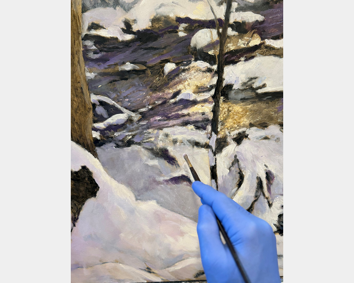 A hand covered in a blue glove holds a small paint brush in front of a painting composed of shades of white, purple, and gold.
