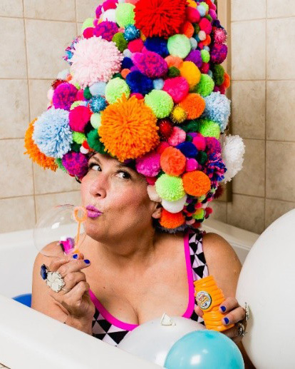 A white woman wearing a big, colorful hat made from puffballs