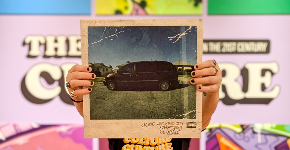A pair of hands hold a Good Kid M.A.A.D City vinyl in front of The Culture title wall