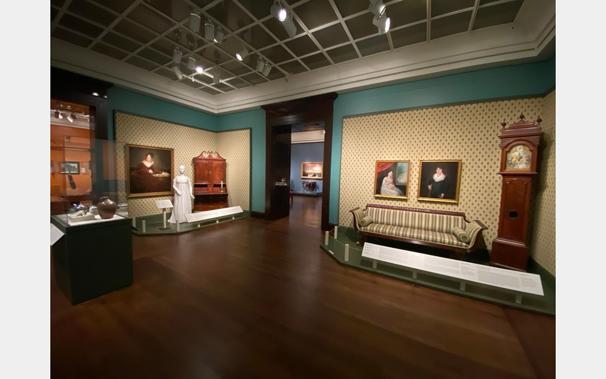 Museum exhibition gallery featuring furniture, paintings, a mannequin in a dress, and a display case.