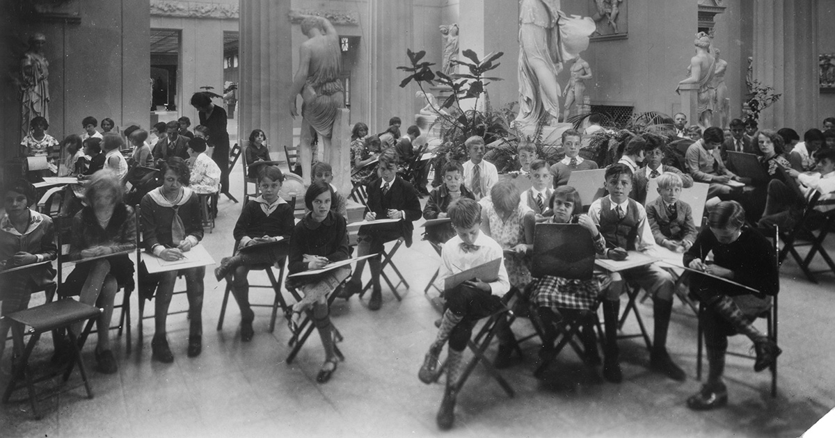 A black and white historical photo of many students sitting in chairs and sketching in a big room with columns
