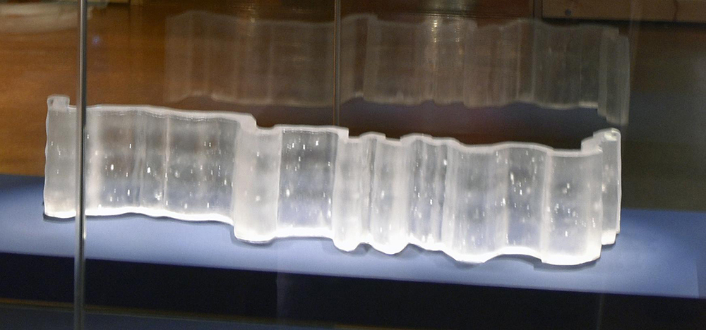 Pa’huk Site is a three-dimensional sculpture crafted from colorless cast glass. This artwork appears as a broad and crooked rectangular band, with bends and dips throughout, reminiscent of a flowing ribbon or the path of a curving river.